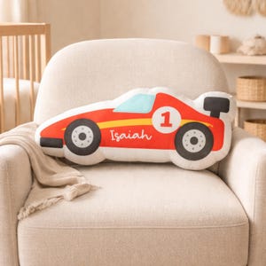 May include: A red race car shaped pillow with black wheels, a blue windshield, and a black spoiler. The number 1 is on the side, and the name "Isaiah" is written in cursive. The pillow is on a beige armchair.