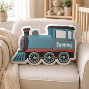 May include: A blue and red train-shaped pillow with the name "Tommy" printed on the side. The pillow has black wheels and a black smokestack. It is placed on a white armchair.