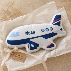May include: A white airplane-shaped pillow with blue and red accents, the name "Noah" is printed on the side. The pillow is presented on white tissue paper with a beige ribbon and a tag that reads "Custom-Made Just for Them".