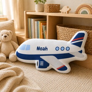 Blue and white airplane shape pillow personalized with child name and photographed on a modern nursery armchair, soft plush aviation themed decorative pillow for boys bedroom decor, custom transportation nursery accessory, travel inspired kids room styling, personalized airplane cushion gift for toddlers, aviation baby room decoration, and playful shaped pillow for cozy nursery and playroom spaces