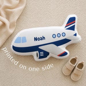 Custom airplane shaped pillow for kids personalized with the name Noah, styled in a soft neutral nursery with modern aviation themed decor, plush airplane cushion for toddler boy bedroom styling, transportation nursery inspiration, pilot baby shower gift ideas, travel themed kids room decoration, cozy personalized plush pillow for reading corners, nursery chairs, beds, and playroom seating areas