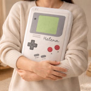 May include: A white and gray handheld video game console pillow with a green screen, a directional pad, red buttons, and the name "Helena" printed on it. The pillow is being held by a person.