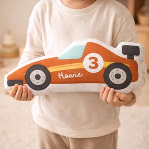 May include: Orange race car shaped pillow with black wheels, a light blue windshield, and a black spoiler. The number 3 is on a white circle, and the name "Howie" is written in white script.