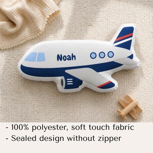 May include: A white airplane-shaped pillow with blue and red accents, the name "Noah" is printed on the side. The pillow has a blue outline and features three blue circles. A small wooden airplane is next to the pillow.