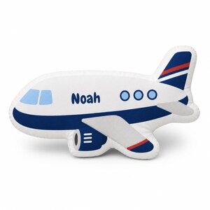 May include: A white airplane-shaped pillow with blue and red accents. The pillow features the name "Noah" in blue lettering, along with blue windows and three blue circles. The wings and tail have blue and red details.