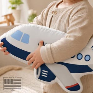 May include: A white airplane-shaped pillow with blue and red accents. The pillow features blue windows and a dark blue stripe along the top. The pillow is being held by a person.