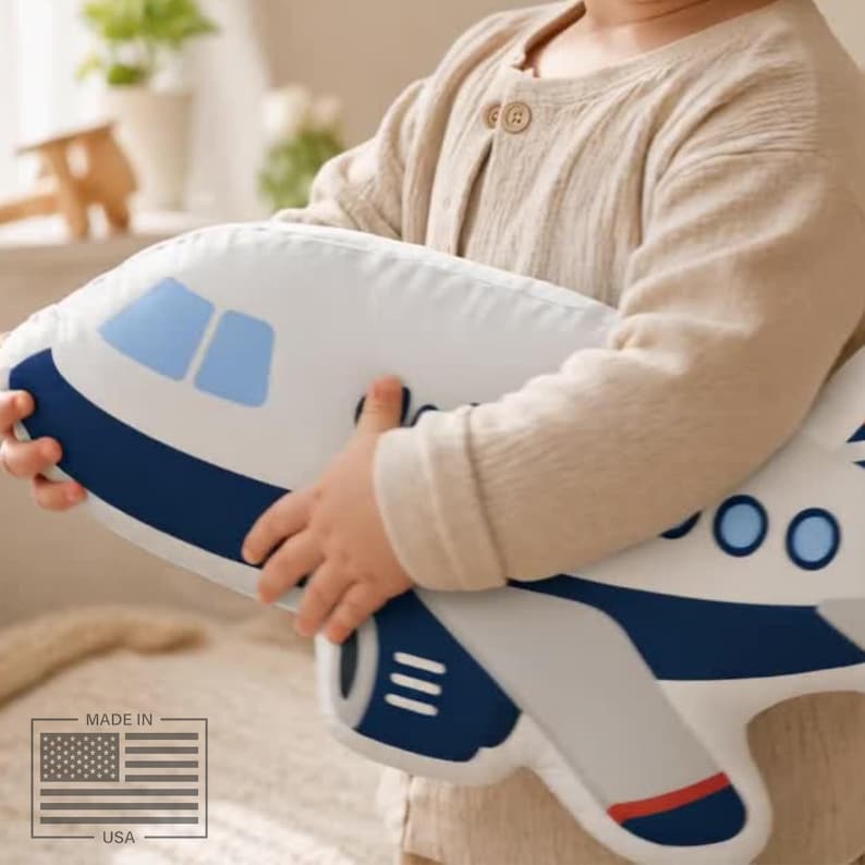 May include: A white airplane-shaped pillow with blue and red accents. The pillow features blue windows and a dark blue stripe along the top. The pillow is being held by a person.