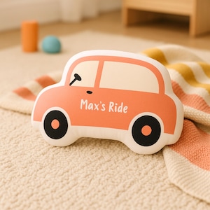 May include: A car-shaped pillow in shades of peach and white, with black wheels and a steering wheel. The text "Max's Ride" is printed on the side. The pillow is on a patterned blanket.