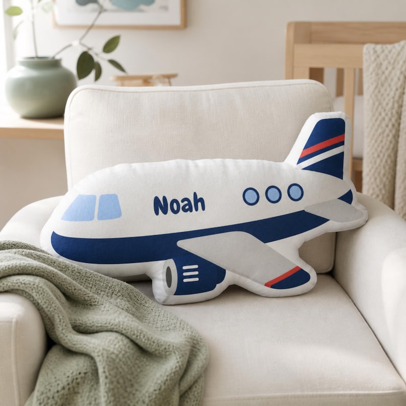 Personalized airplane shape pillow with the name Noah displayed on a cozy cream nursery chair, featuring a blue and white airplane design perfect for aviation nursery decor, toddler boy room styling, transportation themed bedroom ideas, custom airplane plush gifts for kids, pilot family nursery inspiration, travel themed playroom decor, and personalized shaped pillow room accessories for babies and toddlers
