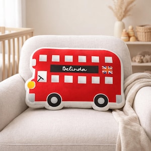 May include: A red London bus-shaped pillow with the name "Belinda" printed on a black rectangle, and a Union Jack flag. The pillow has white windows, black wheels, and a yellow headlight. It is placed on a white armchair.