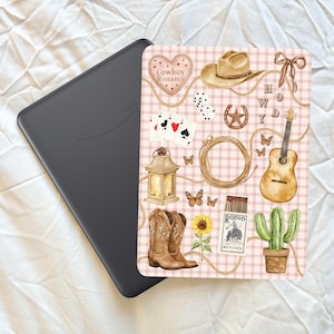 May include: A black e-reader next to a decorative cover with a cowboy theme. The cover features a heart with "Cowboy Romance" text, a cowboy hat, guitar, boots, and other western-themed illustrations on a pink plaid background.