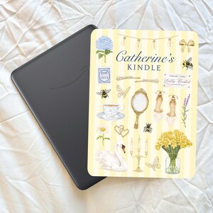 May include: A black Kindle e-reader next to a decorative card with a yellow striped background. The card features illustrations of flowers, bees, a swan, and the text "Catherine's Kindle".