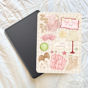 May include: A black e-reader next to a rectangular sticker sheet featuring watercolor illustrations of cozy items including a pink blanket, a lamp, slippers, pajamas, socks, a sleep mask with the text "NAP TIME," and signs reading "let's stay in bed" and "Shhh. I'm Reading!"