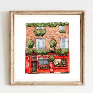 May include: Watercolor painting of The Temple Bar pub in Dublin, Ireland, framed in a wooden frame. The pub's red facade contrasts with the brick building and green foliage. The sign reads "The Temple Bar Estd 1840".