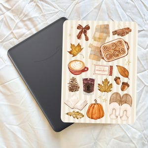 May include: A black e-reader device next to a rectangular sticker featuring watercolour illustrations of autumn items including a brown bow, plaid scarf, cinnamon sticks, cinnamon rolls, coffee, matches, leaves, candle, book, pumpkin, and slippers.