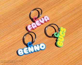 May include: Three colorful keychains with names: "FREYA" in pink, "BENNO" in blue, and "BUCS" in green and yellow. Each keychain has a silver ring. The keychains are on a wooden surface.