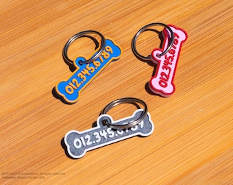 May include: Three dog bone-shaped ID tags with metal rings. The tags are blue, red, and gray with white numbers on them. The numbers on the tags are 012.345.6789.