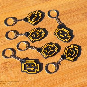 May include: Seven black and yellow keychains with different expressions. The keychains are shaped like a car engine with the word "CHECK" on one keychain.