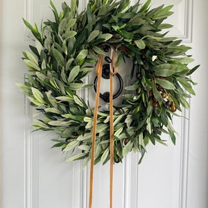 May include: A lush olive wreath with green leaves and dark olives, accented by a brown ribbon. The wreath is hanging on a white door, creating a natural and inviting aesthetic. Perfect for home decor.