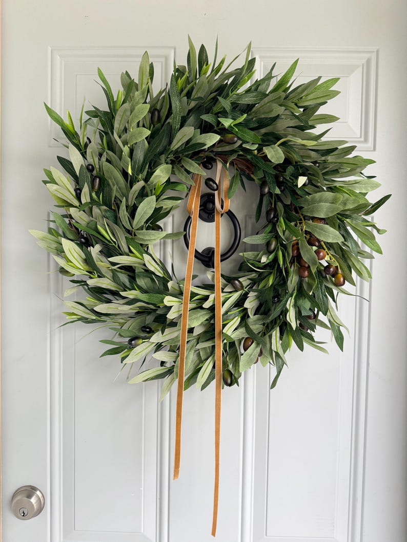 May include: A lush olive wreath with green leaves and dark olives, accented by a brown ribbon. The wreath is hanging on a white door, creating a natural and inviting aesthetic. Perfect for home decor.
