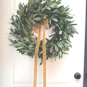 May include: A round olive leaf wreath with a gold ribbon hanging on a white door. The wreath features green leaves and small, dark olives. The gold ribbon is tied in a bow and hangs down. A black doorknob and handle are visible.