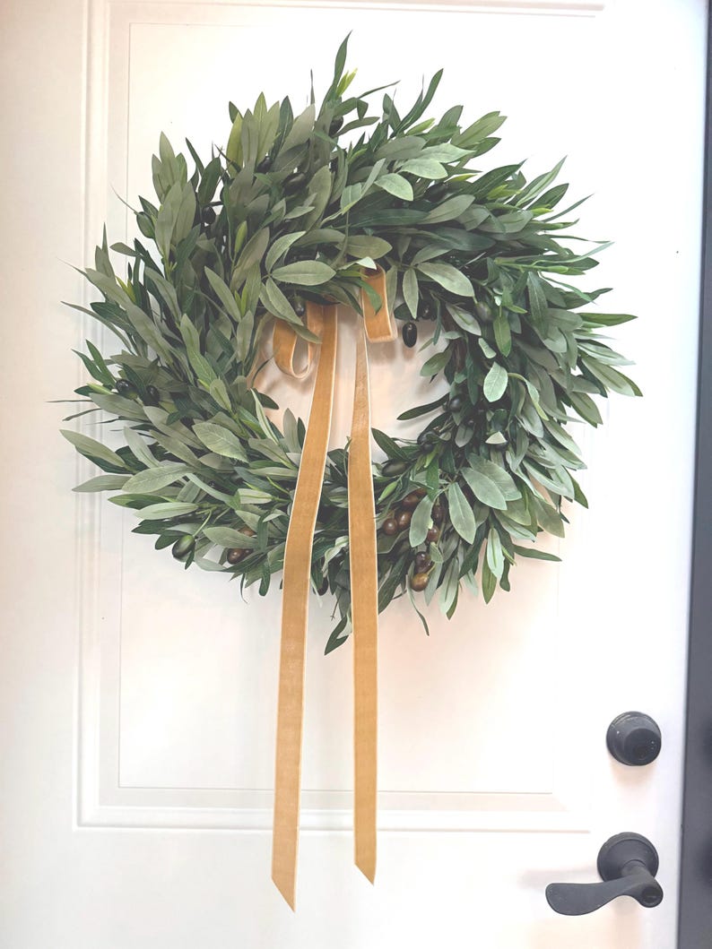May include: A round olive leaf wreath with a gold ribbon hanging on a white door. The wreath features green leaves and small, dark olives. The gold ribbon is tied in a bow and hangs down. A black doorknob and handle are visible.