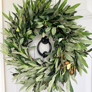 May include: A round olive wreath with green leaves and dark olives. The wreath is hanging on a white door with a black door knocker. The wreath is a decorative item for the front door.