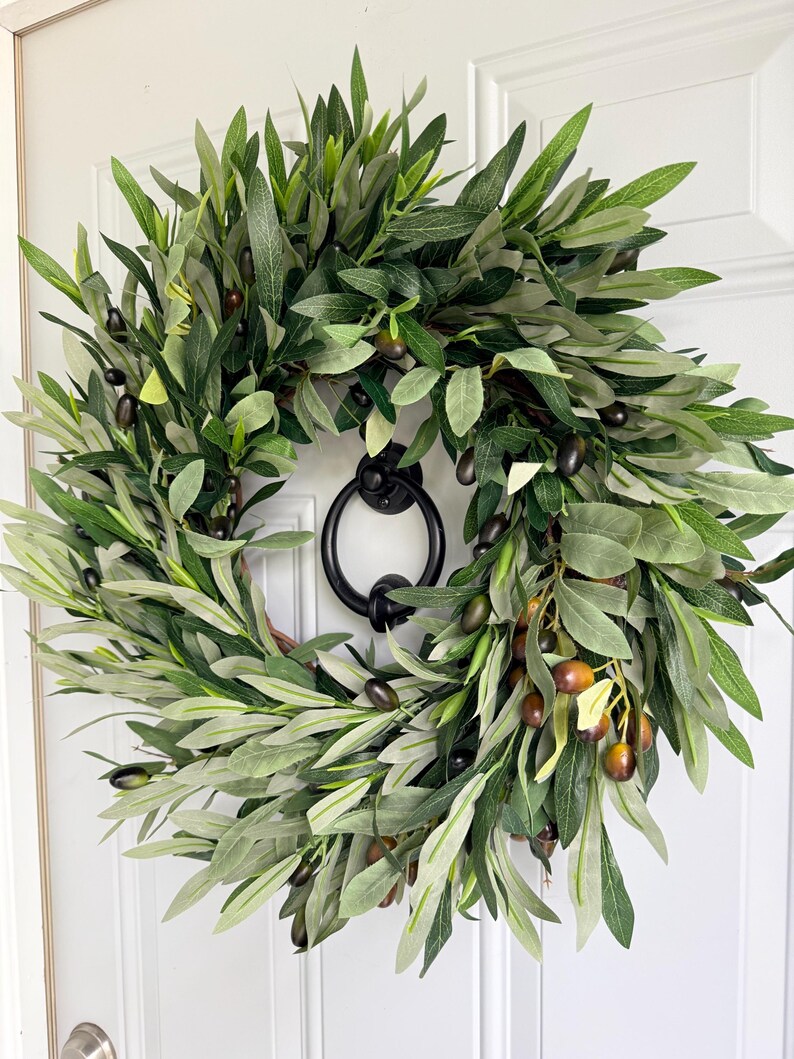 May include: A round olive wreath with green leaves and dark olives. The wreath is hanging on a white door with a black door knocker. The wreath is a decorative item for the front door.