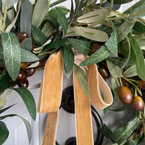 May include: A close-up of an olive wreath with green leaves and brown olives. A gold velvet ribbon is tied in a bow. The wreath is hanging on a white door with a black door knocker.
