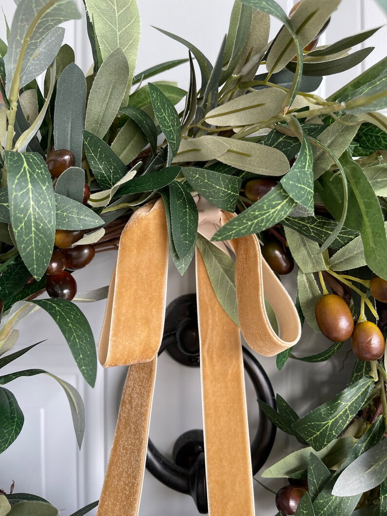 May include: A close-up of an olive wreath with green leaves and brown olives. A gold velvet ribbon is tied in a bow. The wreath is hanging on a white door with a black door knocker.