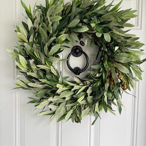 May include: A lush, green olive wreath with dark olives and leaves, hanging on a white door. The wreath has a full, natural appearance, and a black door knocker is centered below it. The door has vertical paneling.