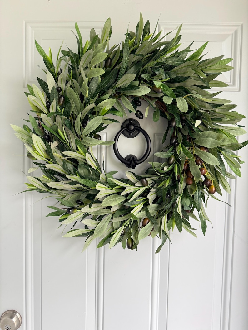 May include: A lush, green olive wreath with dark olives and leaves, hanging on a white door. The wreath has a full, natural appearance, and a black door knocker is centered below it. The door has vertical paneling.
