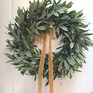 May include: A lush, green olive leaf wreath with dark olives and a gold velvet ribbon. The wreath is a circular design, perfect for adding a touch of nature to any home decor. The leaves are a mix of light and dark green.