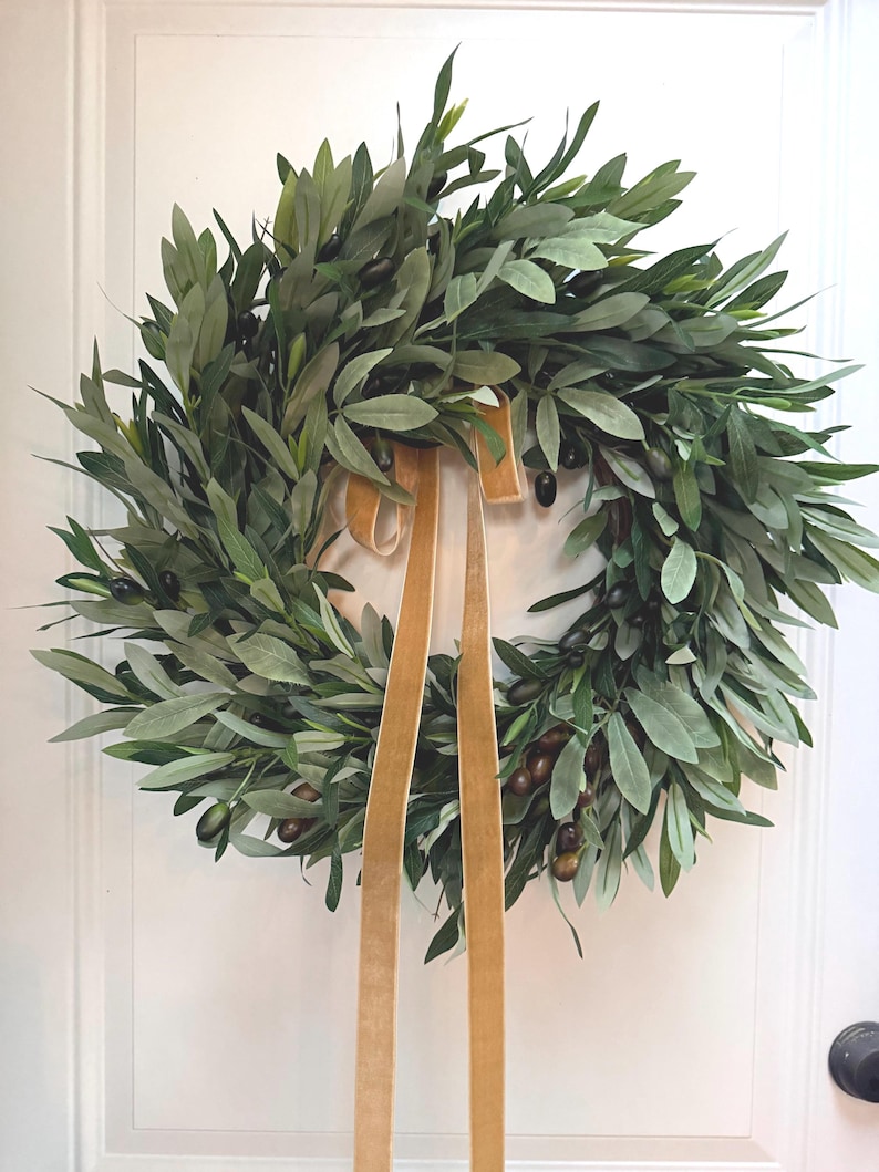 May include: A lush, green olive leaf wreath with dark olives and a gold velvet ribbon. The wreath is a circular design, perfect for adding a touch of nature to any home decor. The leaves are a mix of light and dark green.