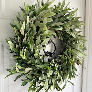 May include: A round olive branch wreath with dark green leaves and black olives. The wreath is hanging on a white door with a black door knocker. The leaves have varying shades of green, creating a natural and textured appearance.