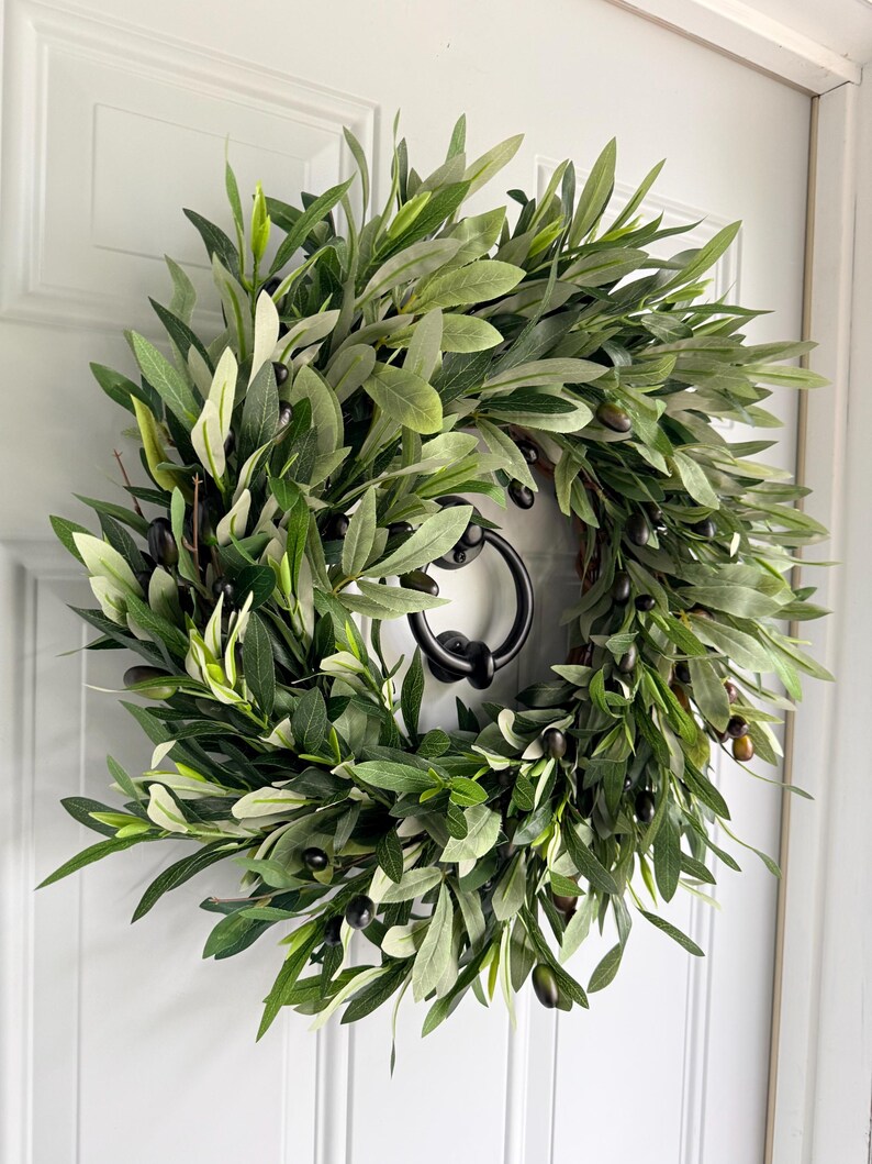 May include: A round olive branch wreath with dark green leaves and black olives. The wreath is hanging on a white door with a black door knocker. The leaves have varying shades of green, creating a natural and textured appearance.