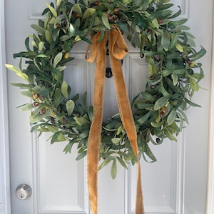 May include: A circular olive wreath with green leaves and dark olives. A tan burlap ribbon is tied in a bow at the top, with long streamers hanging down. The wreath is hanging on a white door.