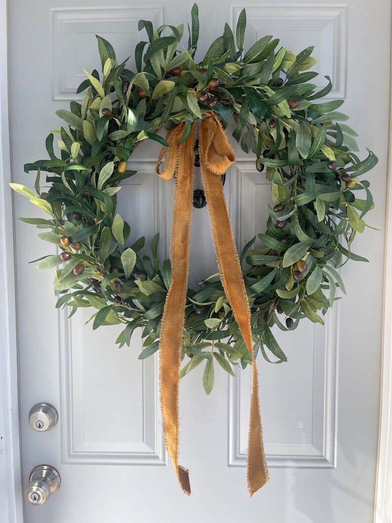 May include: A circular olive wreath with green leaves and dark olives. A tan burlap ribbon is tied in a bow at the top, with long streamers hanging down. The wreath is hanging on a white door.