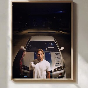 May include: A framed wall art print featuring a person in a white t-shirt standing in front of a silver sports car with blue racing stripes and a rear spoiler. The car has various sponsor decals on the hood and is parked on a dark street at night.