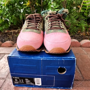 Op de afbeelding: Paar roze en groene sneakers met bruine veters en zolen, op een blauwe doos. De sneakers hebben een suède-achtige textuur op de neus en zijkanten, met een camouflagepatroon aan de bovenkant.