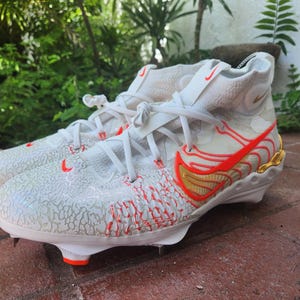 Könnte beinhalten: Weiße und rote Sportschuhe mit einem goldenen Swoosh-Logo. Die Schuhe haben ein strukturiertes Obermaterial und eine weiße Sohle mit roten Akzenten.