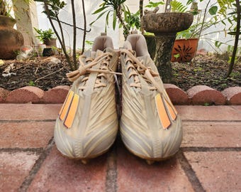 May include: A pair of beige and orange football cleats with a textured design. The cleats have a lace-up closure and a black stripe on the side.