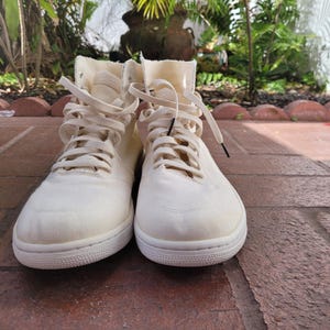 May include: A pair of white high-top canvas trainers with white laces. The shoes have a simple design and are made of a soft, breathable material.