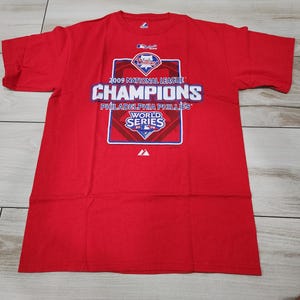 Puede incluir: Camiseta roja con el texto "2009 National League Champions Philadelphia Phillies World Series". La camiseta tiene cuello redondo y mangas cortas. El gráfico es en blanco, azul y rojo.
