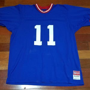 Pode incluir: Uma camisa azul de malha com o número 11 em branco na frente. A camisa tem acabamentos vermelhos e brancos na gola e nas mangas. Uma etiqueta é visível no canto inferior direito.