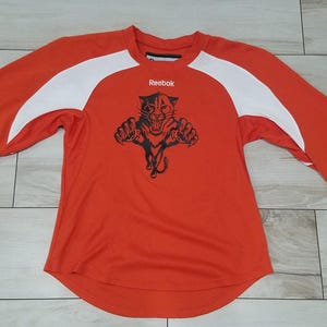 May include: Orange and white long-sleeved shirt with a black panther logo and the word "Reebok" printed on the front.