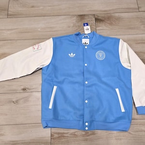 May include: Light blue and white varsity jacket with the Adidas logo on the chest. The jacket has white sleeves, a light blue body, and a button-up front. A patch is visible on the left sleeve. The jacket has a round emblem on the chest.
