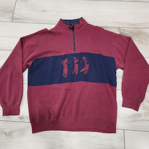 May include: A maroon and navy blue quarter-zip sweatshirt with an embroidered design of four golfers. The sweatshirt has a ribbed collar and cuffs.