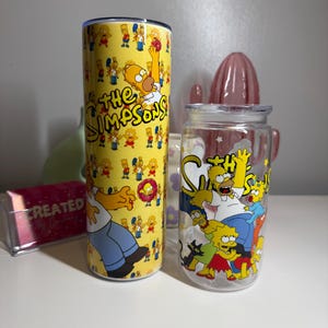 May include: A yellow tumbler and a clear glass jar featuring characters from "The Simpsons." The tumbler has a yellow background with Homer Simpson and other characters. The jar has a lid and features the Simpson family.