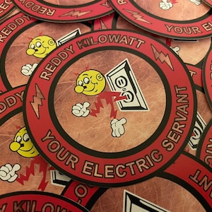 Puede incluir: Una pila de pegatinas circulares rojas y negras con un personaje de dibujos animados que sostiene un rayo y el texto "Reddy Kilowatt Your Electric Servant".